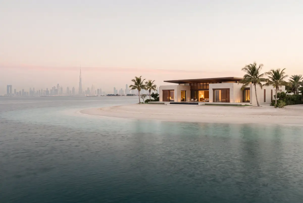 Une villa de plain-pied parmi les palmiers sur une petite île de sable blanc, avec la silhouette de Dubaï à peine visible à l'horizon